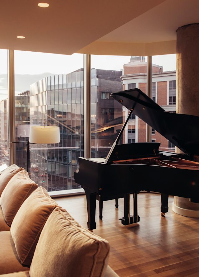 Corner suite of the Conrad Washington. A baby grand piano sits in front of large floor-to-ceiling windows with a view of the city in background.