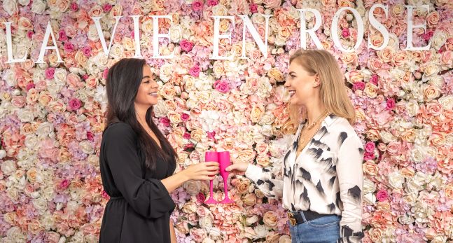 Two woman holding pink champagne glasses standing in front of a wall of pink and white roses