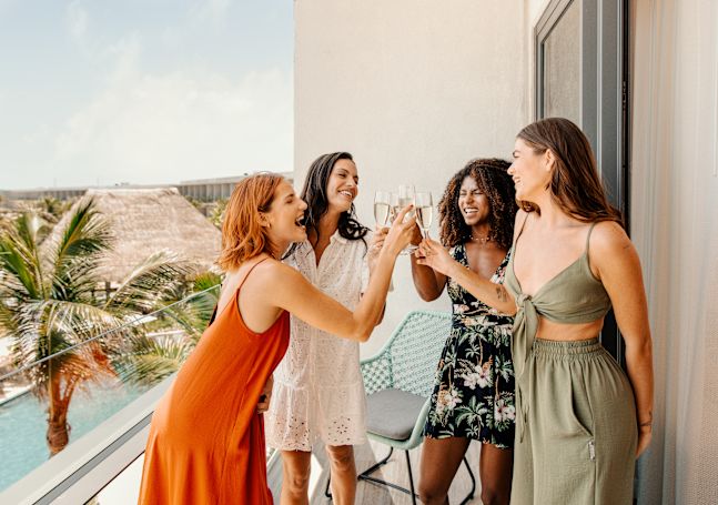 A group of four women raise a drink together on a hotel balcony