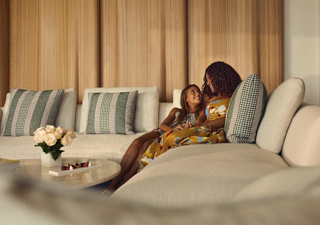 Two people sitting on a curved sofa in a guest room with patterned pillows and a round table with flowers.
