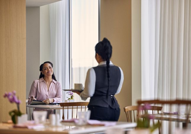 Frau sitzt am Tisch und lächelt beim sich nähernden Mitarbeiter