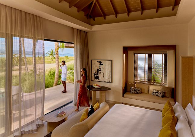 Two people in a sunlit bedroom suite, one standing on the deck looking toward the landscape while the other enjoys a drink near the open sliding doors, with views of palm trees and distant water.