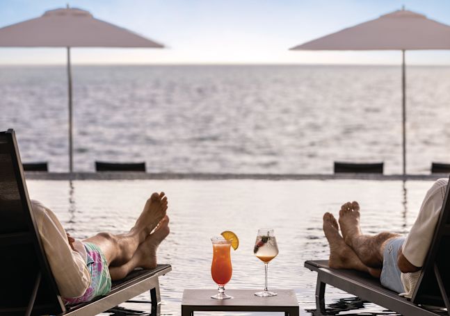 Two people sit in chairs with drinks next to pool by the ocean