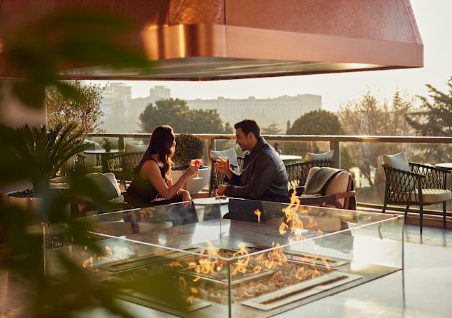 Doi oaspeți stau unul lângă celălalt pe o terasă exterioară a hotelului, ținând în mână băuturi care privesc spre oraș.