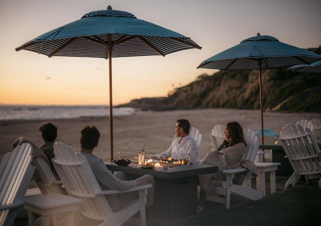 a group of people sitting around a fire pit and having drinks at the beach