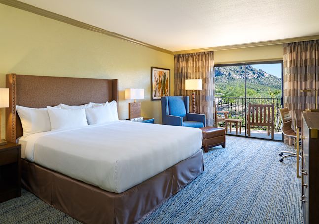 King Bed and Bedside Tables Facing TV and Work Desk with Wall Art and Reading Chair in Corner by Balcony with Mountain View