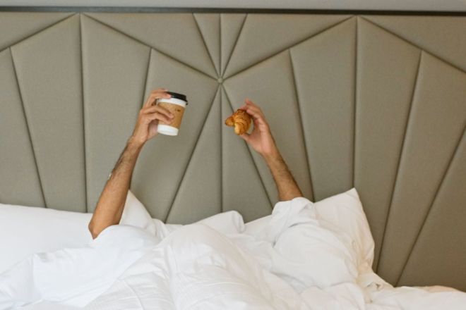 Person Holding Coffee and a Croissant while in Bed in a Hotel Room