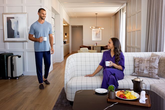 Man walking through guest room towards woman, who is smiling and relaxing on couch