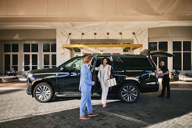 Woman stepping out of car smiling. Her door is being help open while staff member is at the trunk