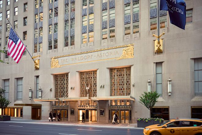 Pintu masuk Waldorf Astoria di Park Avenue dengan kelompok tiga orang yang tiba, beberapa orang berjalan melewatinya, dan seorang anggota tim di pintu utama. Taksi kuning NYC melaju melewatinya.