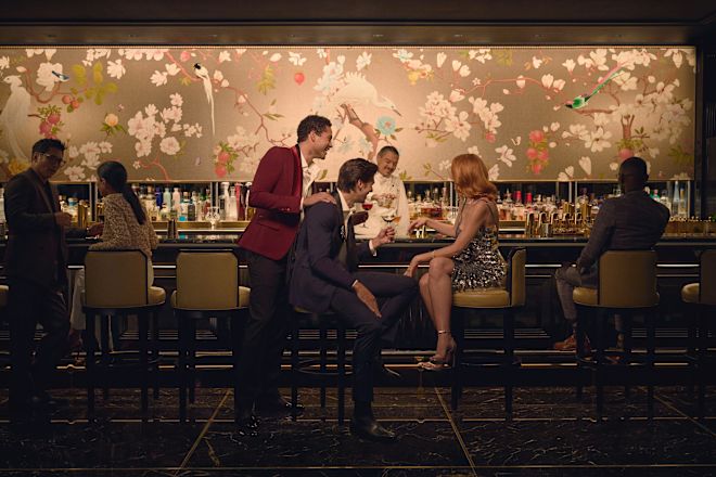 Dua pria dan seorang wanita duduk di bar Peacock Alley sambil berbicara dan mengangkat minuman mereka untuk menghibur. Seorang anggota tim sedang menyiapkan minuman di belakang bar sementara seorang tamu sedang menikmati minuman di sebelah kanan grup dan seorang pria dan wanita sedang mengobrol di sebelah kiri.