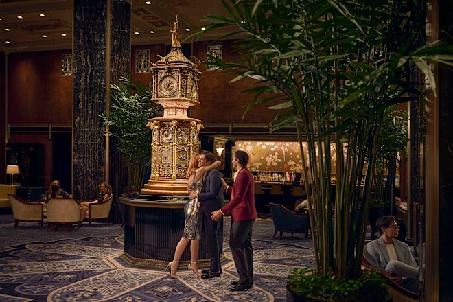 Seorang wanita dengan gaun koktail perak elegan bertemu dua pria dengan setelan malam di Clock in Peacock Alley dan merangkul salah satu pria tersebut.