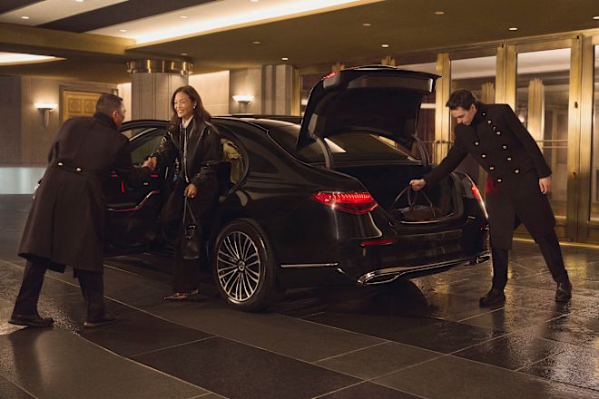 Seorang wanita dengan pakaian berwarna gelap nan elegan tiba di porta dan mengenakan limusin hitam dan disambut oleh anggota tim sementara anggota tim lain membongkar kopernya.