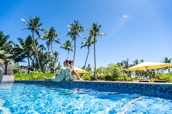 Zwei Personen sitzen am Rande des Swimmingpools mit Palmen, blauem Himmel und Sonnenschirm im Hintergrund