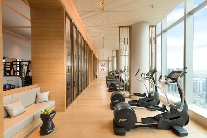 Fitness center with long row of treadmills and ellipticals, facing floor-to-ceiling windows with outdoor view