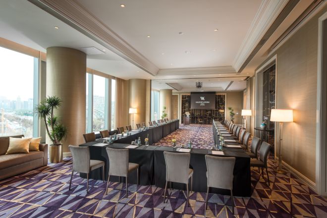Meeting room with long U-shaped tables and chairs facing presentation screen, and floor-to-ceiling windows with outdoor view