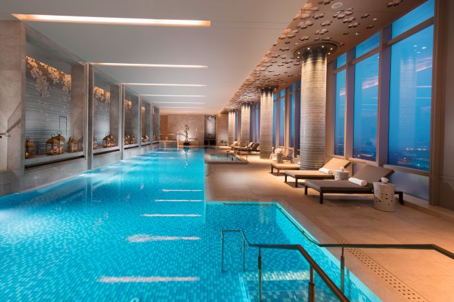 Lavish indoor pool with lounge chairs and floor-to-ceiling windows with outdoor view, lit up at night
