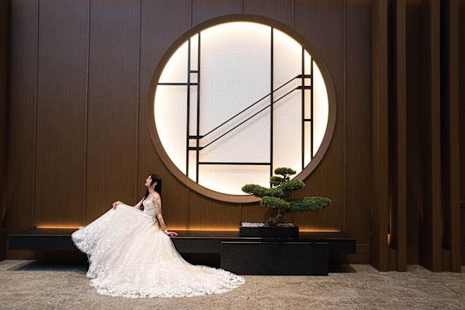 Bride in gown sitting in front of artistic window feature.