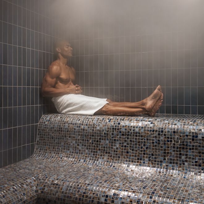Man sitting in the steam room wearing a towel, and with legs out and crossed.