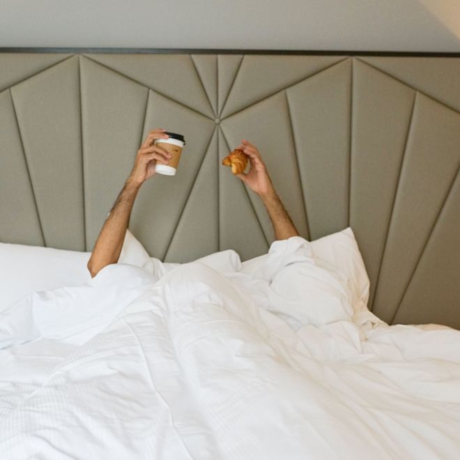Person Holding Coffee and a Croissant while in Bed in a Hotel Room