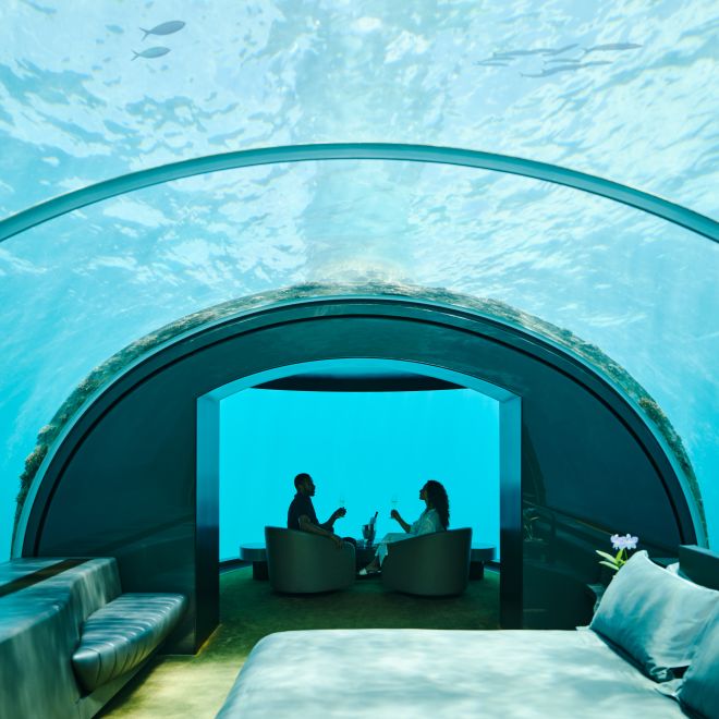 Underwater bedroom, with man and woman relaxing on chairs while fish swim overhead