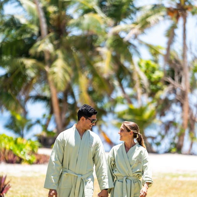 Paar hält Hände, trägt Bademäntel und geht im Hotelgarten mit Palmen im Hintergrund spazieren