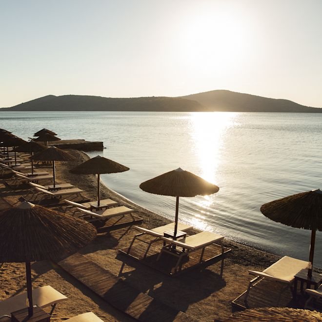Beach with rows of loungers and umbrellas at sunset, with mountains in the distance.