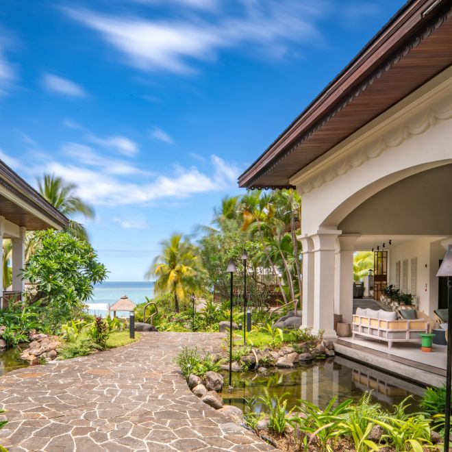 Path between buildings towards beach, with water and trees