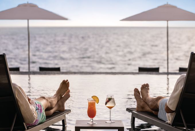Two people sit in chairs with drinks next to pool by the ocean