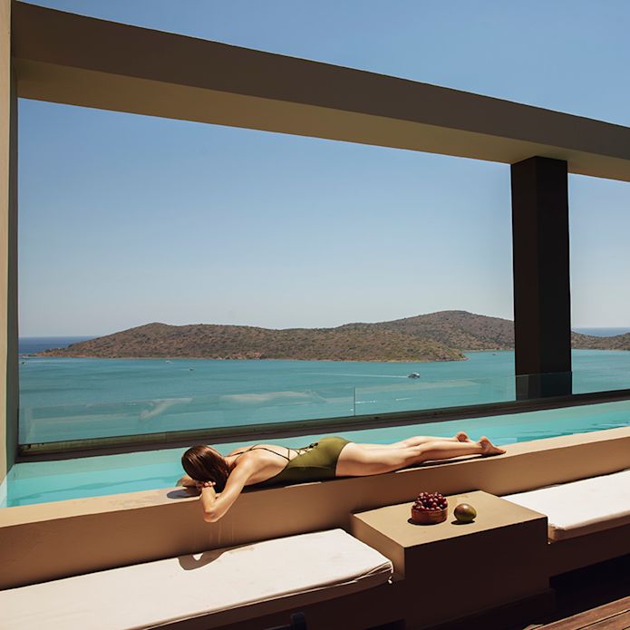 Woman suntanning on a terrace with white cushions on benches, overlooking the sea and hills.