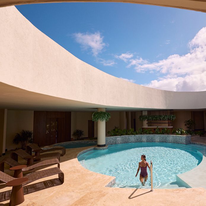Persoană ieșind dintr-o piscină circulară sub un luminator deschis, înconjurată de arhitectură curbată, scaune lounge și cer albastru strălucitor.