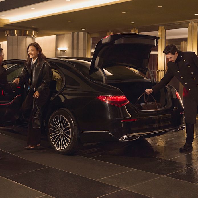 Une femme habillée d'élégants vêtements sombres arrive dans la porte cochère dans une limousine noire et est accueillie par un membre du personnel tandis qu'un autre membre du personnel décharge ses bagages.