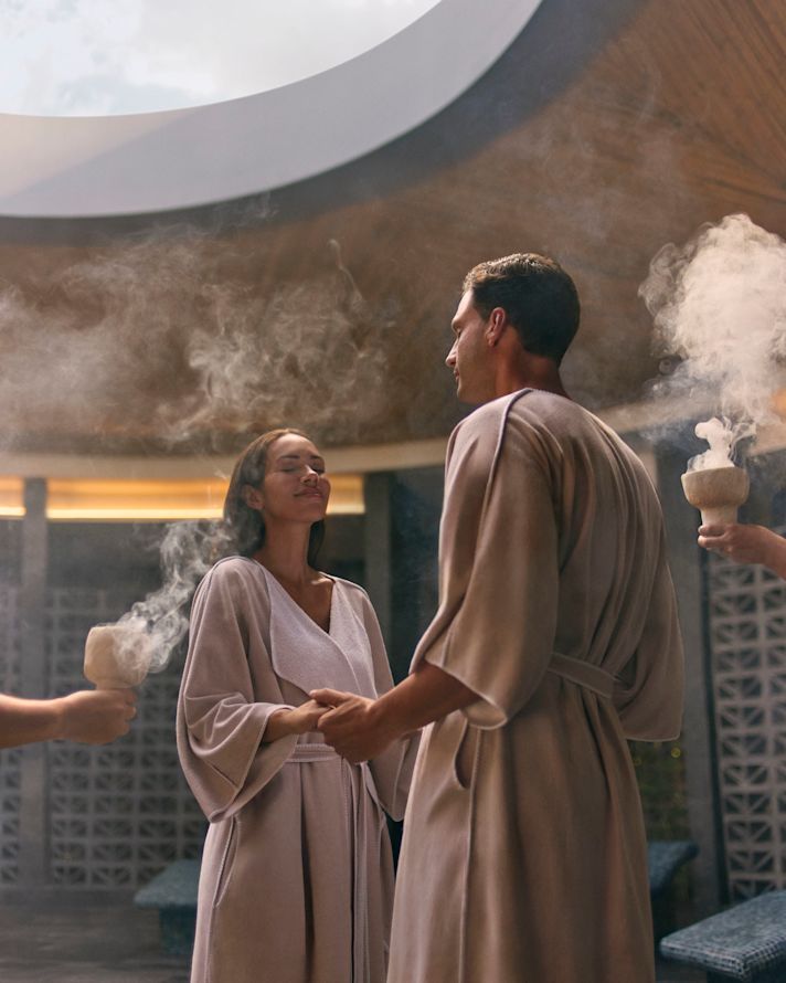 Bir kadın ve bir adam, spada ayakta dururken ellerini tutuyor ve ekip üyeleri sigara içilebilen tütsülü kaseler tutuyor