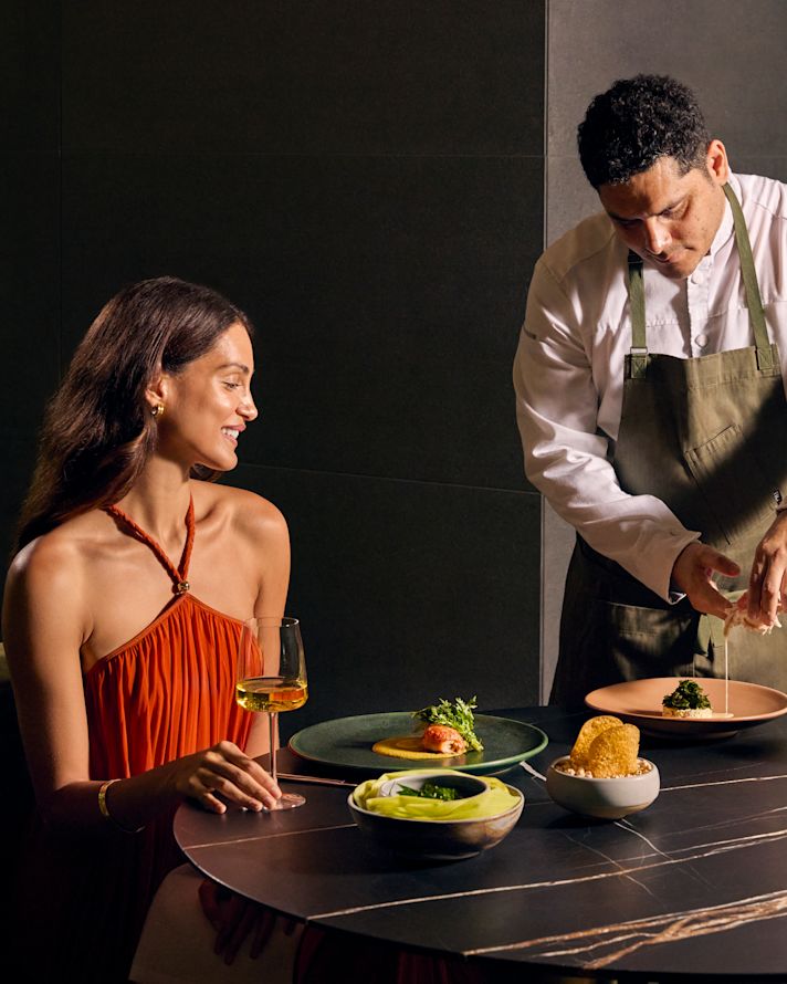 Mulher sorrindo sentada à mesa do restaurante, com um membro da equipe servindo comida