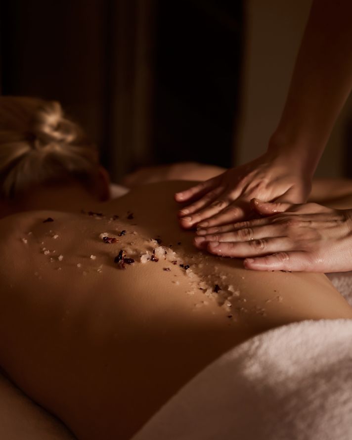 Imagem em close das costas de uma mulher recebendo uma massagem com grãos de sal e flores