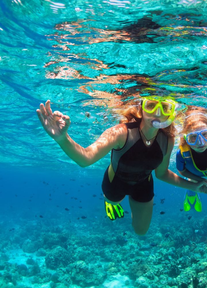 Madre y niño practicando esnórquel con máscara, bucean bajo el agua con peces tropicales en la piscina del arrecife de coral. Muestra con las manos la señal de buceo O