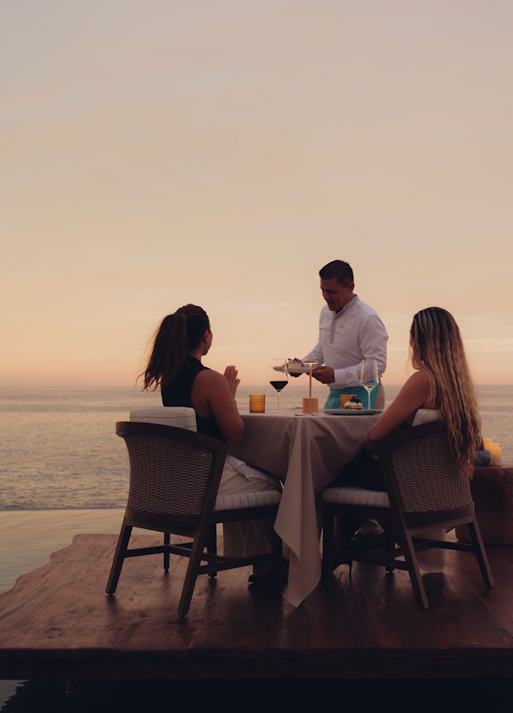 Oceanfront dining table with two guests and a team member at sunset.