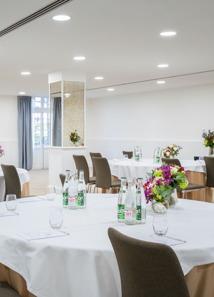 Sala de conferință La Fontaine cu mese rotunde în lenjerie albă, fiecare are o vază din sticlă transparentă plină de flori strălucitoare și apă îmbuteliată.