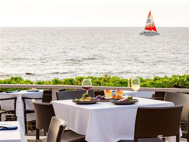 A outdoor table with dinner served, facing the ocean. A sailboat floats in the distance.