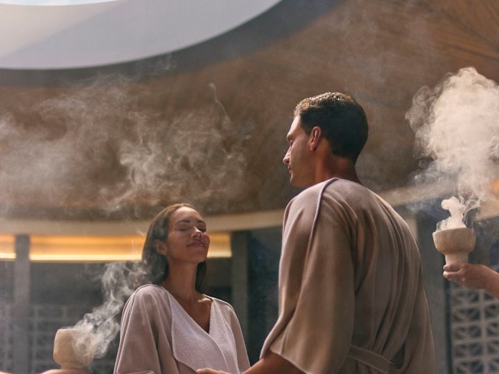 A woman and a man are holding hands while standing in the spa and  team members are holding bowls with smoking incense