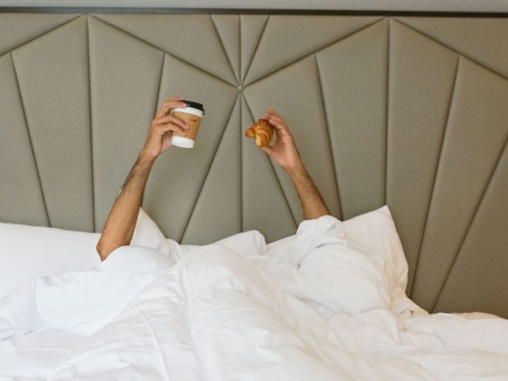Person Holding Coffee and a Croissant while in Bed in a Hotel Room