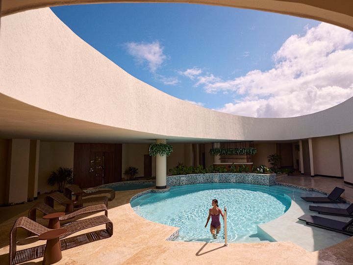 Person stepping out of a circular pool beneath an open skylight, surrounded by curved architecture, lounge chairs, and bright blue sky.