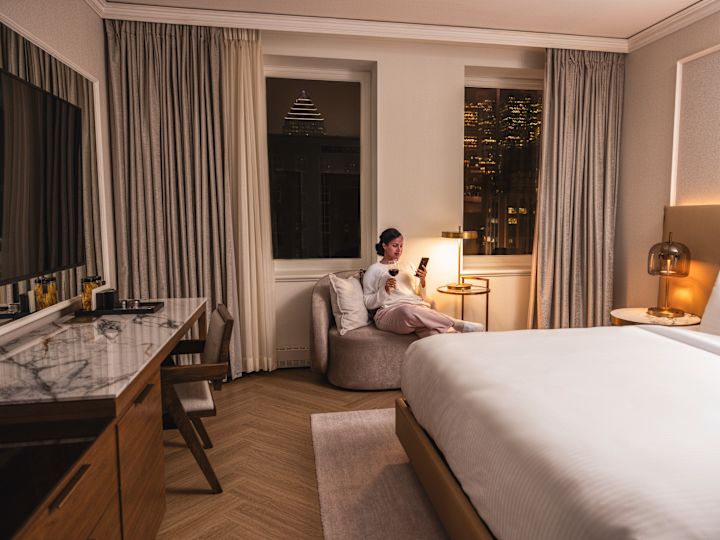 Bed in guest room with woman relaxing in comfortable chair with drink