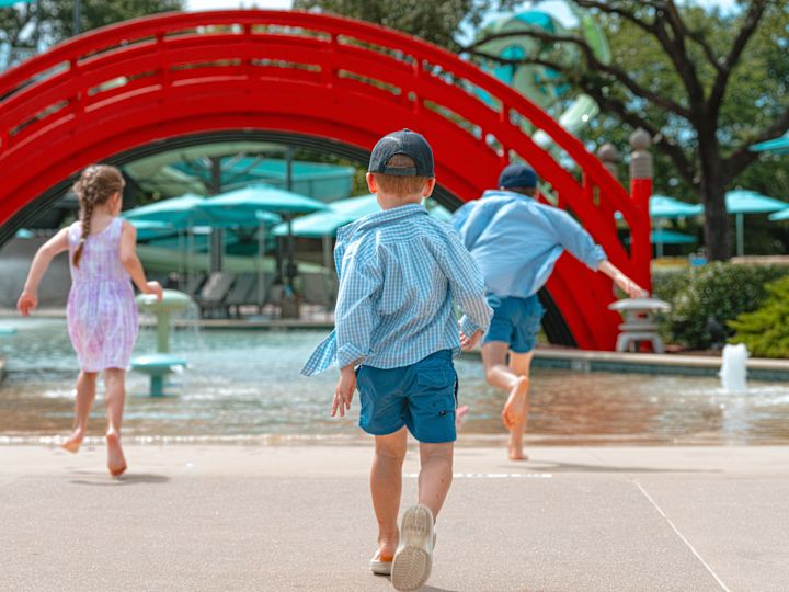 Niños corriendo en el parque acuático Jade Waters hacia una fuente de agua espumosa y un puente rojo