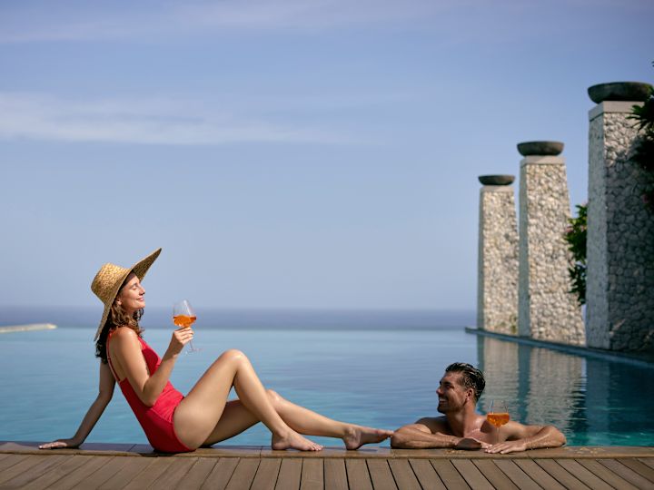 Man in pool, with arms resting on side as woman reclines with drink in her hand