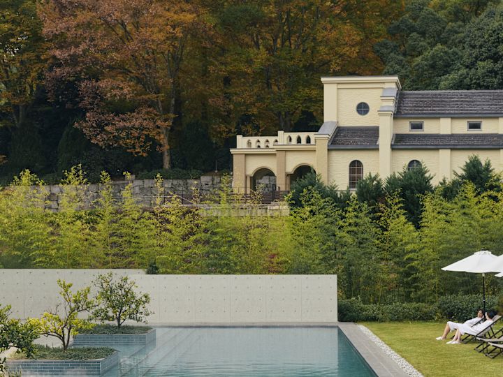 Persona che nuota in una piscina esterna circondata da vegetazione, con sedie a sdraio e ombrelloni sul prato e alberi autunnali dietro un edificio color crema.