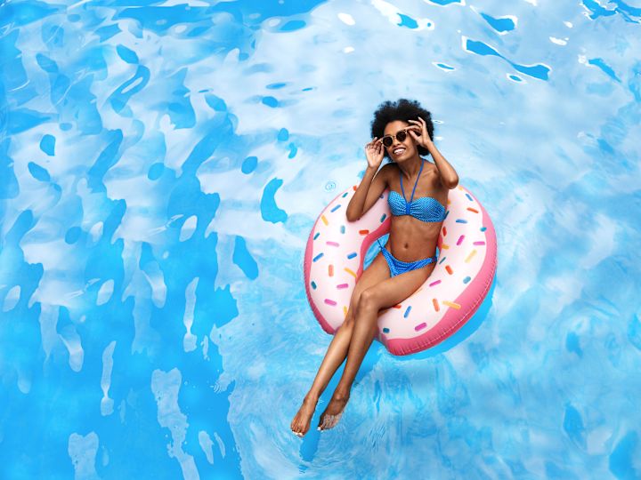 Schwarze Frau in blauem Bikini, die im tropischen Urlaub auf einem aufblasbaren Ring am Swimmingpool schwimmt.