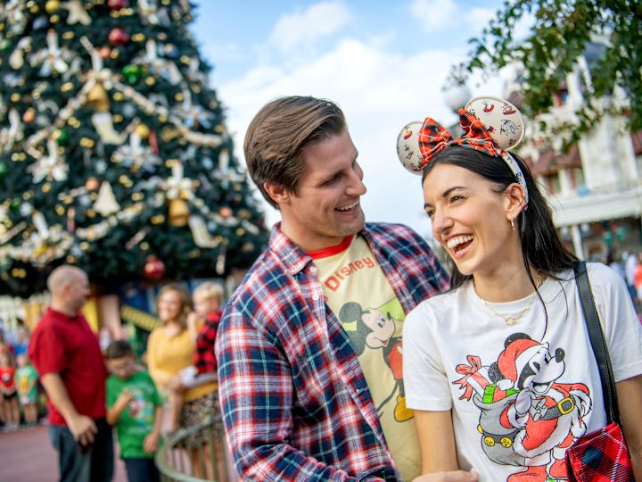 Couple smiling with Disney themed t-shirts and ears and Christmas tree in the background.