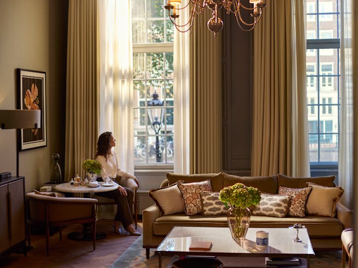 Elegant suite with a sofa accented by patterned cushions, a coffee table with flowers, and tall windows framed by cream curtains, with a person seated at a small dining table.