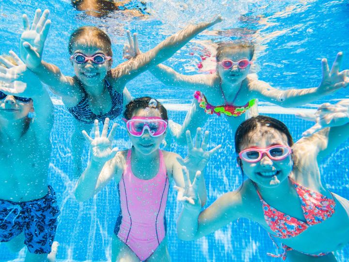 Kids wearing goggles underwater in a pool waving at the camera